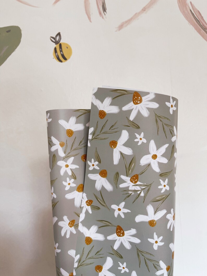 Op de afbeelding: Twee rollen grijs inpakpapier met een wit madeliefje bloemenpatroon. Het papier heeft een subtiel groen en geel kleurenschema.