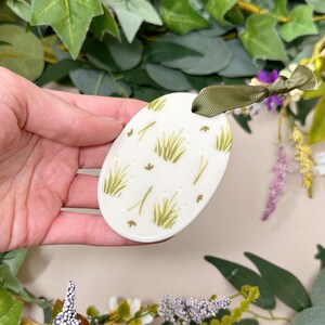 Op de afbeelding: Een wit ovaalvormig keramiek ornament met een groen gras patroon. Het ornament heeft een groen lint aan de bovenkant.