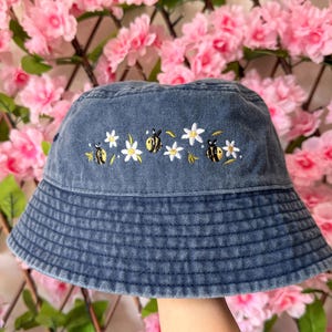May include: A denim bucket hat with embroidered bees and white flowers. The hat is a light blue color and has a structured brim. The embroidery is in yellow, black, and white. The hat is a casual accessory.