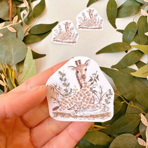 May include: A sticker featuring a hand-drawn illustration of a baby giraffe sitting in a patch of green grass. The giraffe is brown and white with a long neck and large ears. The sticker is on a white background.