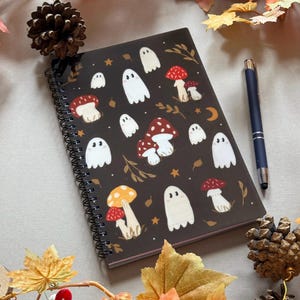 May include: A dark brown spiral-bound notebook decorated with white ghost and mushroom illustrations. A blue pen lies next to the notebook, surrounded by autumn leaves and pine cones. The design includes stars and a crescent moon.
