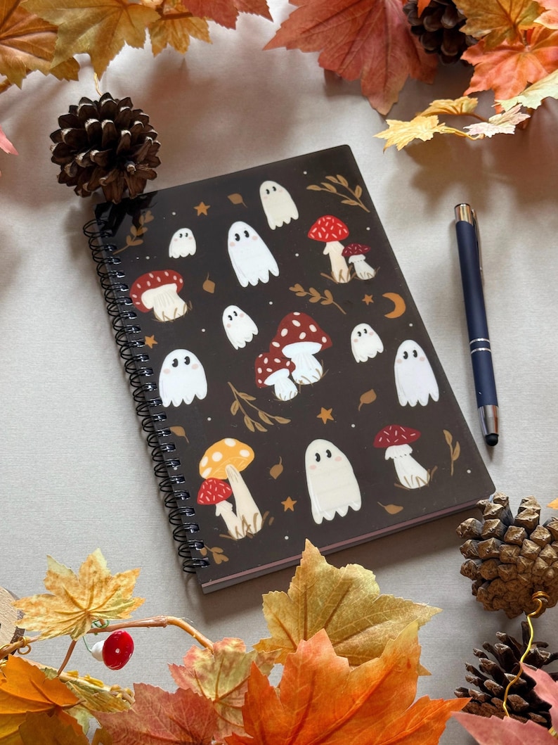 May include: A dark brown spiral-bound notebook decorated with white ghost and mushroom illustrations. A blue pen lies next to the notebook, surrounded by autumn leaves and pine cones. The design includes stars and a crescent moon.