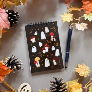 May include: A dark brown spiral-bound notebook with illustrations of white ghosts and red mushrooms. A dark blue pen lies beside the notebook, surrounded by autumn leaves, pine cones, and a wooden jack-o'-lantern.