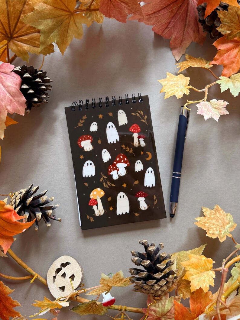 May include: A dark brown spiral-bound notebook with illustrations of white ghosts and red mushrooms. A dark blue pen lies beside the notebook, surrounded by autumn leaves, pine cones, and a wooden jack-o'-lantern.