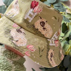 May include: A green bucket hat with a repeating pattern of sheep, tulips, and white picket fences. The hat has a white brim and is made of a soft fabric. The overall theme is a pastoral, springtime design.