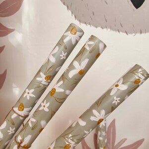 Op de afbeelding: Drie rollen groen inpakpapier met een wit madeliefje bloemenpatroon. Het papier is vastgebonden met touw en heeft een rustieke, landelijke uitstraling.