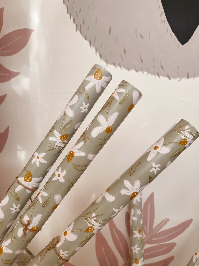 Op de afbeelding: Drie rollen groen inpakpapier met een wit madeliefje bloemenpatroon. Het papier is vastgebonden met touw en heeft een rustieke, landelijke uitstraling.
