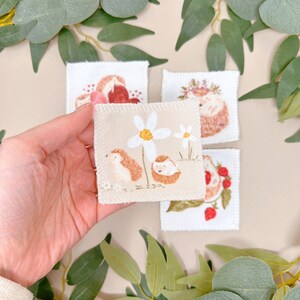 Puede incluir: Cuatro cuadrados de tela bordados con un dise&ntilde;o de erizo. Un cuadrado muestra un erizo con una corona de flores, otro muestra un erizo con un coraz&oacute;n, otro muestra un erizo con una flor y el &uacute;ltimo muestra un erizo con fresas.