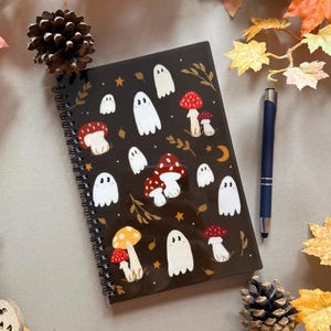 May include: A black spiral-bound notebook decorated with white ghost and mushroom illustrations. A blue pen lies next to the notebook, with autumn leaves and pine cones arranged around it.