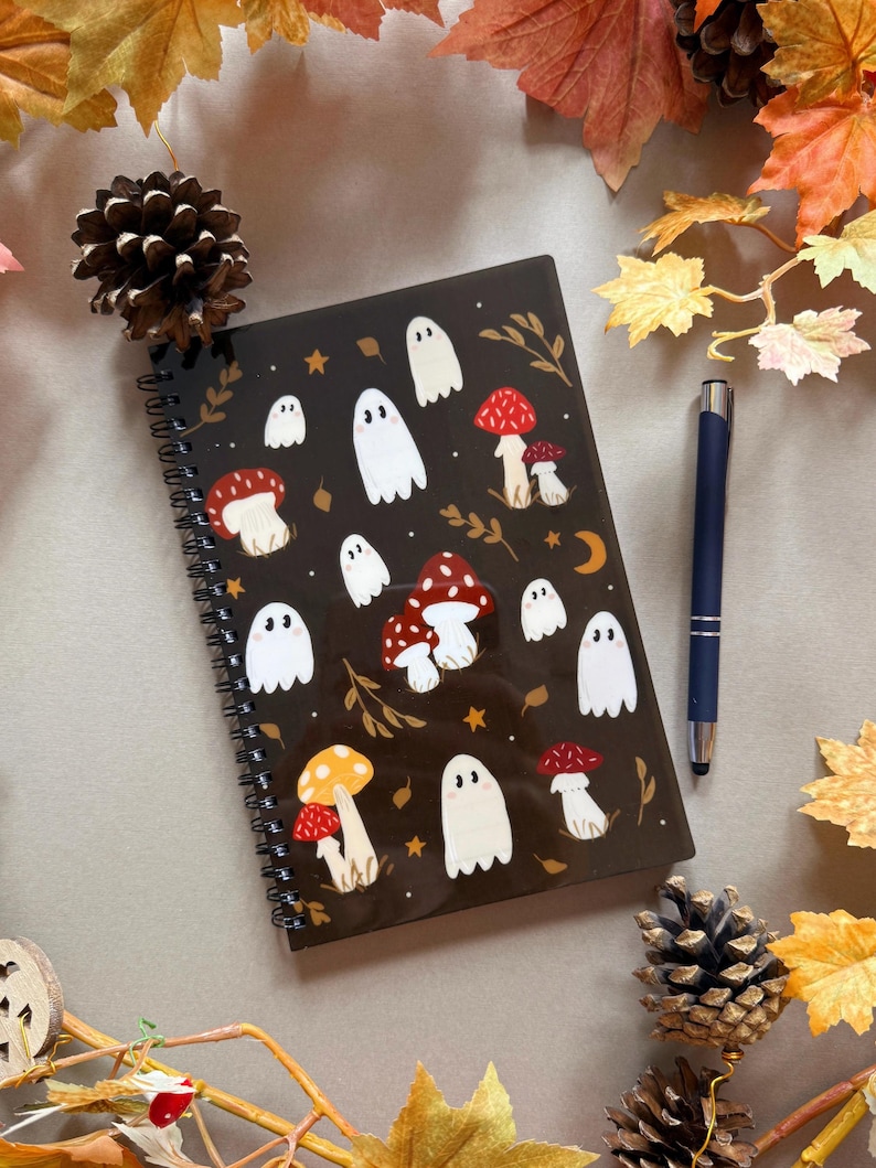 May include: A black spiral-bound notebook decorated with white ghost and mushroom illustrations. A blue pen lies next to the notebook, with autumn leaves and pine cones arranged around it.