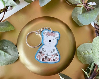 Op de afbeelding: Een sleutelhanger in de vorm van een witte beer met een bloemenpatroon en een blauwe omtrek. De beer draagt een bloemenkroon en is bevestigd aan een zilveren ketting.
