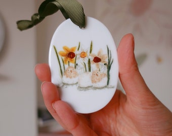 Pode incluir: Ornamento oval de cerâmica branca com um design pintado à mão de três coelhos brancos cercados por narcisos amarelos e brancos com caules verdes.