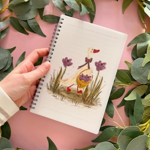 May include: A white spiral bound notebook featuring a hand-drawn illustration of a duck carrying a basket of purple flowers. The duck is wearing a pink bow and has red feet.