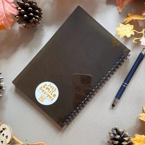 May include: A dark brown spiral-bound notebook with a sticker reading "Just Smile Designs" and a QR code. A blue pen lies next to the notebook. The image is decorated with autumn leaves and pine cones.