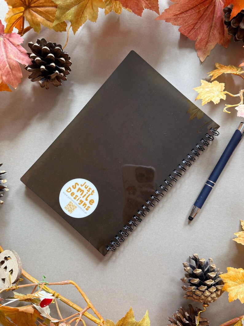 May include: A dark brown spiral-bound notebook with a sticker reading "Just Smile Designs" and a QR code. A blue pen lies next to the notebook. The image is decorated with autumn leaves and pine cones.