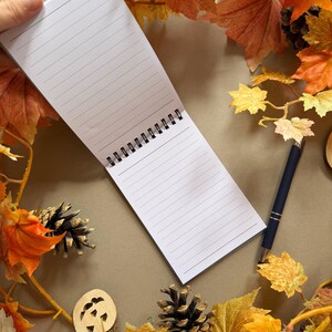 May include: A lined notepad, held open, surrounded by autumnal leaves in shades of orange, yellow, and brown. A dark blue pen lies beside the notepad. Decorative pine cones and a wooden pumpkin accent the scene, creating a fall theme.