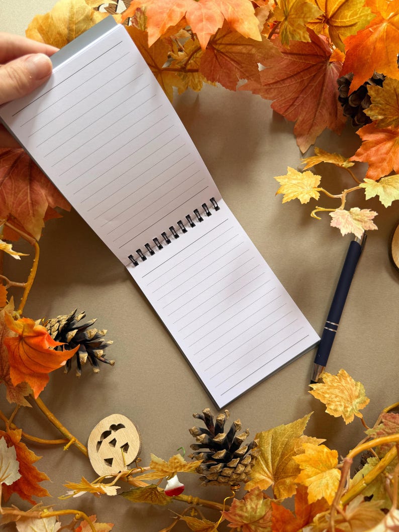 May include: A lined notepad, held open, surrounded by autumnal leaves in shades of orange, yellow, and brown. A dark blue pen lies beside the notepad. Decorative pine cones and a wooden pumpkin accent the scene, creating a fall theme.