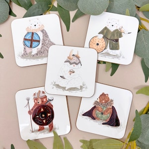 Może przedstawiać: Zestaw czterech kwadratowych podkładek pod kubki z ilustracjami niedźwiedzi w różnych strojach inspirowanych wikingami. Każda podkładka ma białe tło z brązową ramką i przedstawia innego niedźwiedzia w hełmie, tarczy i innych dodatkach. Wszystkie niedźwiedzie są białe, z wyjątkiem jednego, który jest brązowy.