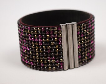 May include: Wide cuff bracelet with a black interior and a surface covered in small, sparkling pink, black, and gold-toned crystals. The bracelet has a silver-colored magnetic clasp.
