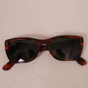 Puede incluir: Gafas de sol marrones con estampado de carey y lentes oscuras. Las gafas tienen una forma de montura rectangular cl&aacute;sica. El logotipo de Ray-Ban es visible en la lente superior izquierda.