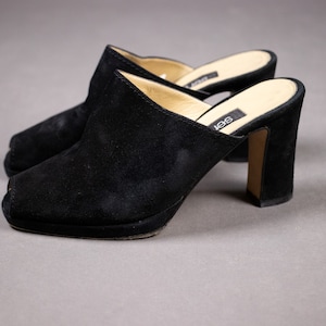 Puede incluir: Un par de mules de tacón de ante negro con puntera cuadrada. Los tacones miden aproximadamente 10 cm de alto.