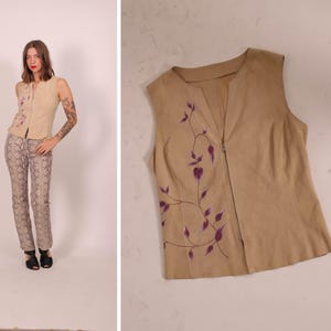 May include: A tan sleeveless vest with a floral design in purple. The vest has a V-neck and a button closure. The model is wearing snake print pants and black shoes.