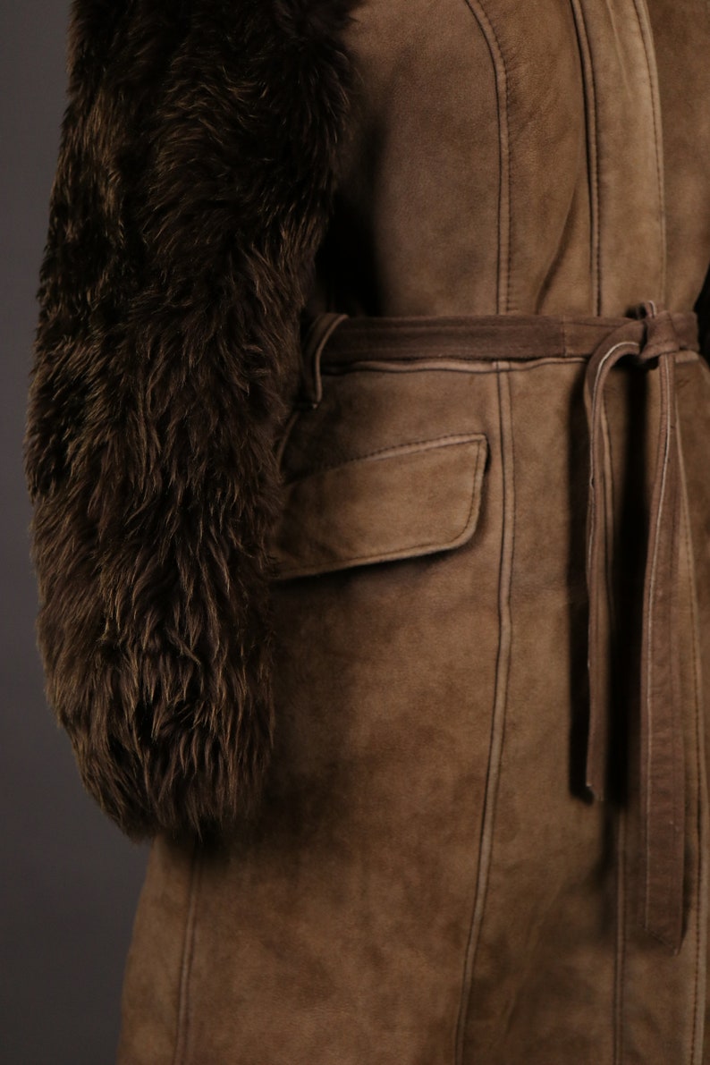 May include: A brown suede jacket with a fur collar and a tie belt. The jacket has a patch pocket on the front and is lined with a brown fabric.