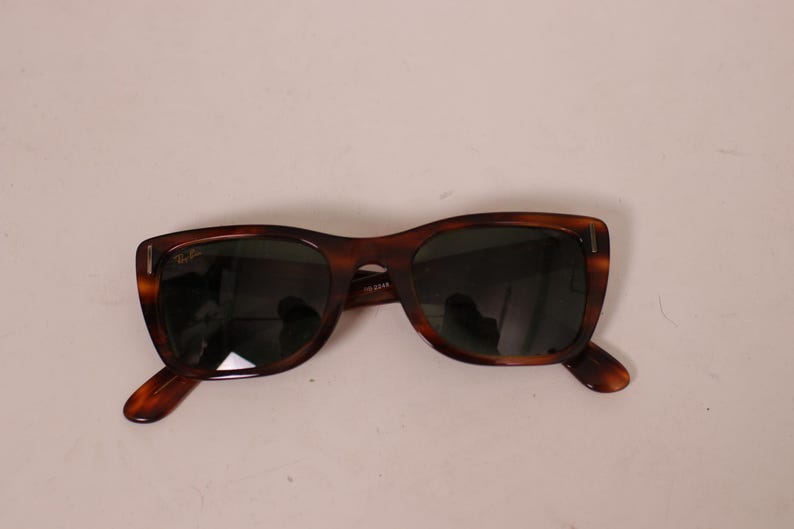 Puede incluir: Gafas de sol marrones con estampado de carey y lentes oscuras. Las gafas tienen una forma de montura rectangular cl&aacute;sica. El logotipo de Ray-Ban es visible en la lente superior izquierda.