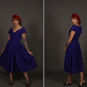 Puede incluir: Una mujer con un vestido morado con un amplio escote de hombros descubiertos y una boina roja. Está de pie frente a un fondo gris.