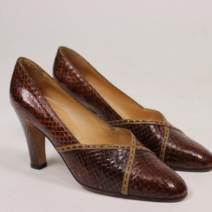 K&ouml;nnte beinhalten: Ein Paar braune Lederschuhe mit hohen Abs&auml;tzen, Schlangenhautmuster und braunem Lederbesatz. Die Schuhe haben eine spitze Zehenpartie und einen geschlossenen R&uuml;cken.