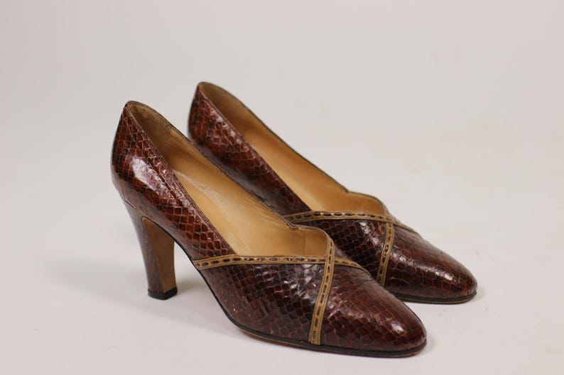 K&ouml;nnte beinhalten: Ein Paar braune Lederschuhe mit hohen Abs&auml;tzen, Schlangenhautmuster und braunem Lederbesatz. Die Schuhe haben eine spitze Zehenpartie und einen geschlossenen R&uuml;cken.
