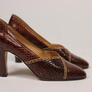 K&ouml;nnte beinhalten: Ein Paar braune Pumps aus Schlangenleder mit braunem Besatz. Die Schuhe haben eine spitze Zehenpartie und einen hohen Absatz.