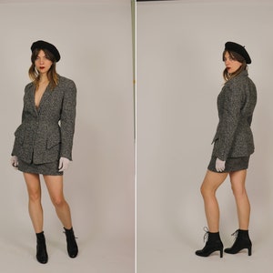 Puede incluir: Un traje de falda de tweed gris con una boina negra. El traje incluye una chaqueta con un solo botón de cierre y una falda de cintura alta. La modelo lleva botines negros de tobillo.