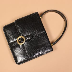 May include: A black handbag with a textured snakeskin pattern, featuring a square silhouette, a single arched handle, and a front flap closure secured by a circular gold-toned metal buckle. The bag has a structured design with visible stitching.