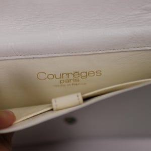 K&ouml;nnte beinhalten: Offene Ansicht einer wei&szlig;en Courr&egrave;ges Paris Handtasche aus Leder. Der Innenraum ist cremefarben mit dem Markennamen in goldfarbenen Buchstaben. Die Tasche ist offen und zeigt eine Tasche und das Innenfutter.