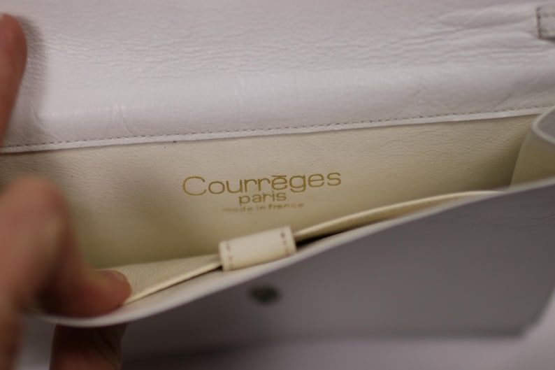 K&ouml;nnte beinhalten: Offene Ansicht einer wei&szlig;en Courr&egrave;ges Paris Handtasche aus Leder. Der Innenraum ist cremefarben mit dem Markennamen in goldfarbenen Buchstaben. Die Tasche ist offen und zeigt eine Tasche und das Innenfutter.