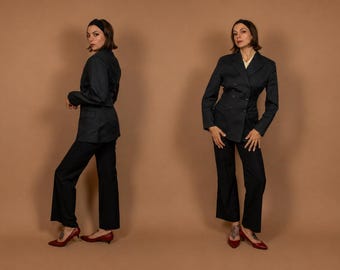 May include: A dark charcoal grey double-breasted blazer with matching straight-leg trousers. The blazer features a notched lapel, long sleeves, and a button-front closure. The outfit is paired with dark red pointed-toe low-heeled shoes.