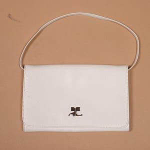 K&ouml;nnte beinhalten: Eine cremefarbene Schultertasche aus Leder mit Klappenverschluss und d&uuml;nnem Riemen. Die Tasche hat ein silbernes Logo auf der Vorderseite. Die Tasche befindet sich vor einem neutralen Hintergrund.