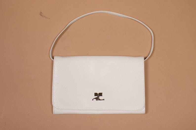 K&ouml;nnte beinhalten: Eine cremefarbene Schultertasche aus Leder mit Klappenverschluss und d&uuml;nnem Riemen. Die Tasche hat ein silbernes Logo auf der Vorderseite. Die Tasche befindet sich vor einem neutralen Hintergrund.