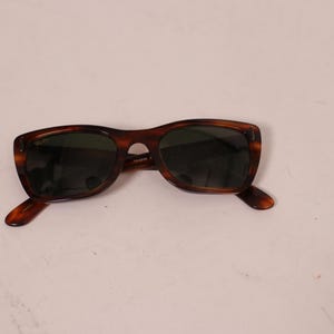 Puede incluir: Gafas de sol marrones con montura de carey y lentes oscuras. Las gafas tienen una montura rectangular cl&aacute;sica. Las patillas de las gafas tambi&eacute;n son de carey. Sobre un fondo blanco.