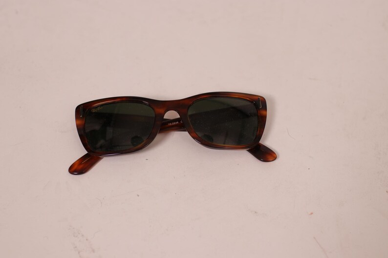 Puede incluir: Gafas de sol marrones con montura de carey y lentes oscuras. Las gafas tienen una montura rectangular cl&aacute;sica. Las patillas de las gafas tambi&eacute;n son de carey. Sobre un fondo blanco.