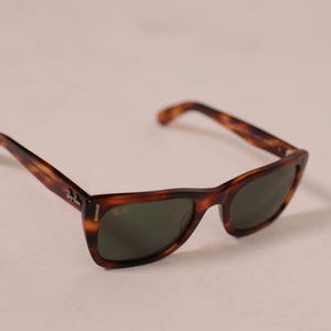 Puede incluir: Gafas de sol de carey con lentes verde oscuro. Las monturas tienen un dise&ntilde;o cl&aacute;sico con un patr&oacute;n marr&oacute;n y &aacute;mbar. El logotipo de Ray-Ban es visible en la patilla.