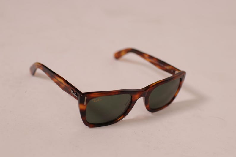 Puede incluir: Gafas de sol de carey con lentes verde oscuro. Las monturas tienen un dise&ntilde;o cl&aacute;sico con un patr&oacute;n marr&oacute;n y &aacute;mbar. El logotipo de Ray-Ban es visible en la patilla.