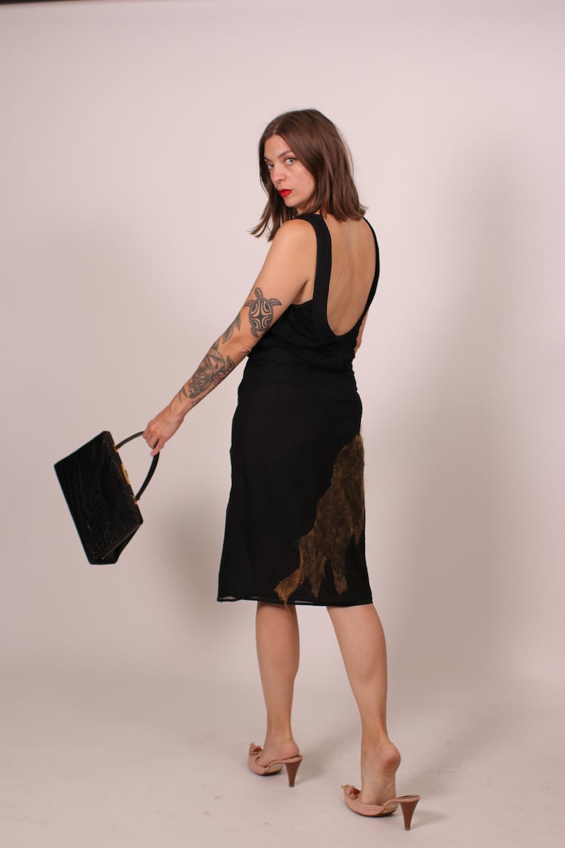 May include: A black dress with a low back and a knee-length skirt. The dress features a brown, textured design on the side. A black handbag with a handle is held in the hand. The person is wearing pink heeled sandals.
