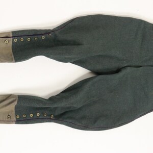 May include: A pair of dark green riding breeches with brown leather trim and button closures. The breeches have a purple seam down the outside of each leg.