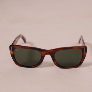 Puede incluir: Gafas de sol marrones con montura de carey y lentes verdes. Las gafas tienen un dise&ntilde;o cl&aacute;sico y retro con una forma ligeramente de ojo de gato. El logotipo de Ray-Ban es visible en la montura.