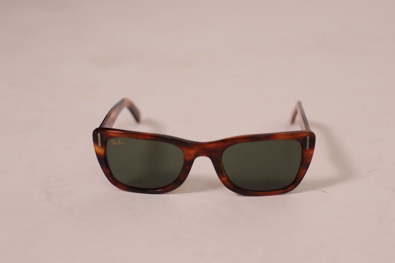 Puede incluir: Gafas de sol marrones con montura de carey y lentes verdes. Las gafas tienen un dise&ntilde;o cl&aacute;sico y retro con una forma ligeramente de ojo de gato. El logotipo de Ray-Ban es visible en la montura.
