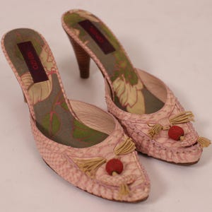 Puede incluir: Un par de mules rosas de tacón alto y punta abierta. Los zapatos tienen un estampado de piel de serpiente, tacones de madera y borlas decorativas con detalles rojos. El interior presenta un diseño floral y el nombre de la marca.
