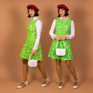 K&ouml;nnte beinhalten: Ein &auml;rmelloses, limettengr&uuml;nes Kleid mit wei&szlig;em Blumenmuster &uuml;ber einem wei&szlig;en lang&auml;rmeligen Hemd mit Kragen. Das Outfit umfasst ein rotes Barett, eine kleine wei&szlig;e strukturierte Handtasche mit Henkel und wei&szlig;e Mary Jane Schuhe mit Blockabsatz.