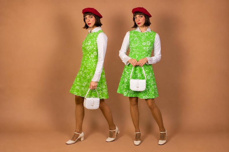 K&ouml;nnte beinhalten: Ein &auml;rmelloses, limettengr&uuml;nes Kleid mit wei&szlig;em Blumenmuster &uuml;ber einem wei&szlig;en lang&auml;rmeligen Hemd mit Kragen. Das Outfit umfasst ein rotes Barett, eine kleine wei&szlig;e strukturierte Handtasche mit Henkel und wei&szlig;e Mary Jane Schuhe mit Blockabsatz.
